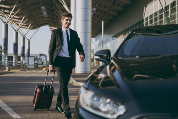 Full body bottom view young traveler businessman young man in black dinner suit going walk outside at international airport terminal with suitcase to car booking taxi Air flight business trip concept.